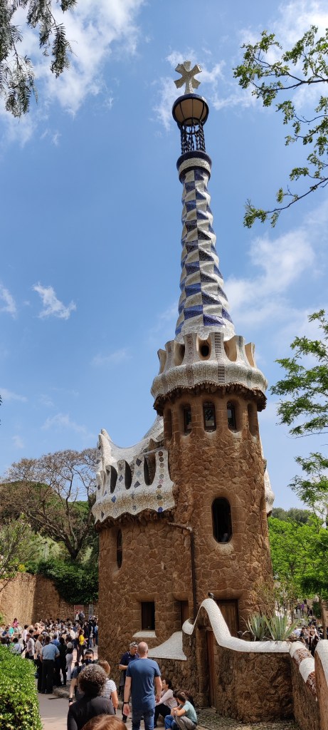 Kapea, ruskea rakennus, jonka katto näyttää piparkakkutalon sokerikuorrutteelta valkosinisine mosaiikkeineen. Ikkuna-aukot ovat osin pyöreitä. Taustalla näkyy paljon ihmisiä, puustoa ja kirkas taivas.