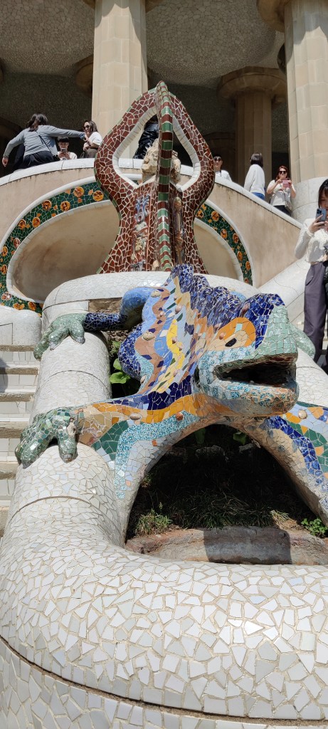 Lähikuva erivärisillä mosaiikkipaloilla koristellusta liskopatsaasta, jolla on suu auki. Se katsoo kohti kameraa ja seisoo raajat levällään vesialtaan päällä, kuin pitäisi kiinni sen reunoista. Taustalla portaikossa näkyy poseeraavia ihmisiä ja lisää mosaiikein koristeltuja muotoja.