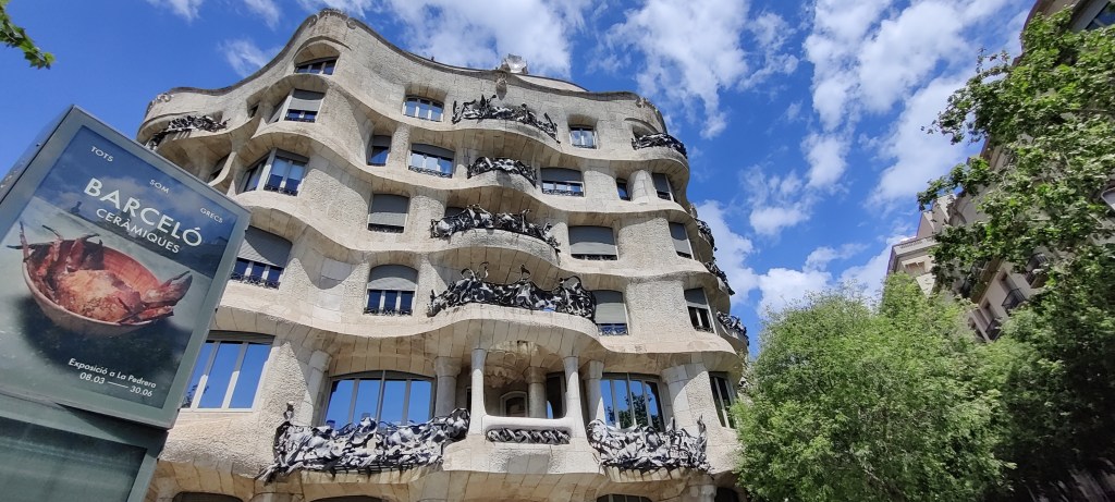 Kuvassa vaalea kerrostalo, jonka ulkoseinät näyttävät aaltoilevan. Parvekkeilla on röpelömäisiä, rautaisia koristeitä. Vasemalla vihreää puustoa, taustalla sininen taivas ja jonkin verran pilviä. Rakennuksen kulmaa peittää mainoskyltti.