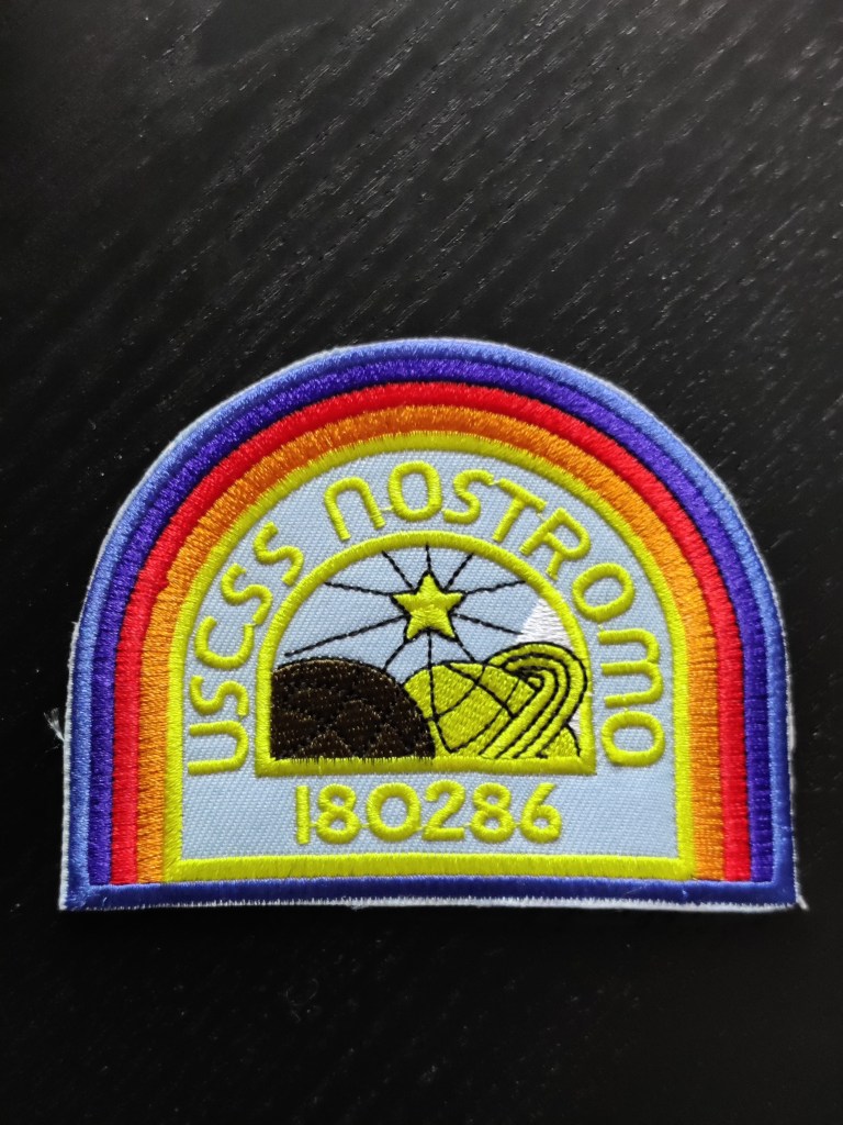Värikäs puoliympyrän mallinen hihamerkki, jossa lukee teksti USCSS NOSTROMO 180286. Hihamerkin keskellä on logo, jossa kaksi planeettaa kohtaa toisensa ja niiden yllä loistaa tähti.