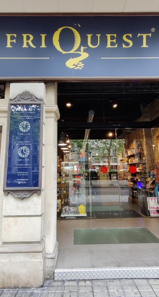 Friquest-nimisen kaupan ulko-ovi. Lasiovien läpi näkyy sisälle kauppaan. Ikkunalasien heijastuksesta huolimatta etualalla näkyy iso keltainen legopää.
Nimikyltin Q-kirjaimessa on polku, jota pitkin kävelee viittaan pukeutunut henkilö.