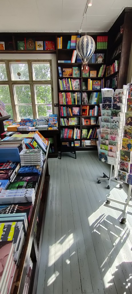 Sisäkuva kirjakaupasta: vasemmalla on matala pöytä täynnä kirjoja. Oikealla etualalla on postikorttitelineitä. Takaseinällä on iso ruutuikkuna, jonka edessä on matala kirjahylly ja jonka vasemmalla puolella on korkea musta kirjahylly, jossa on värikkäitä lastenkirjoja.
Lattia on valkeaksi maalattua puuta, katosta roikkuu pieni lelukuumailmapallo.