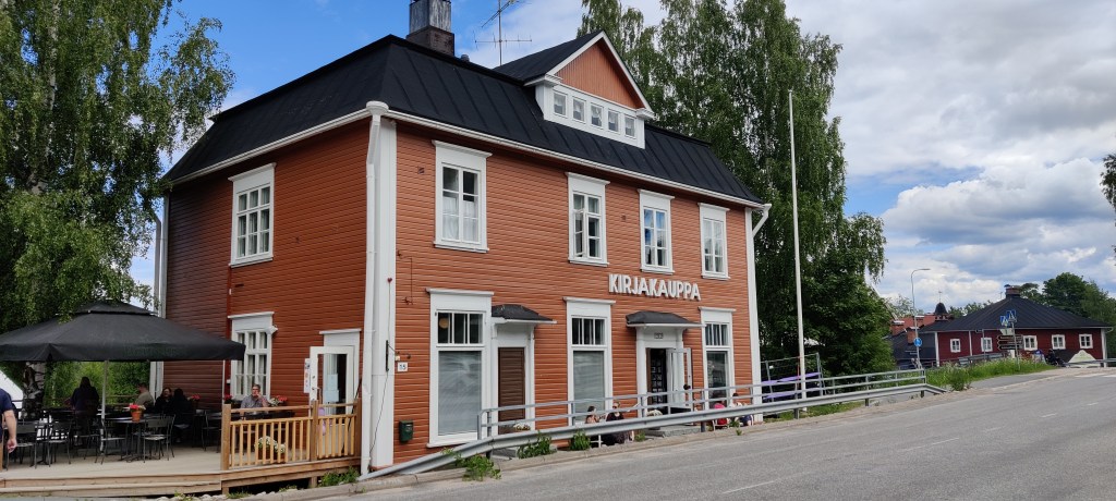 Iso, kaksikerroksinen puutalo seisoo tien vieressä. Talo on oranssi ja sen lukuisissa ikkunoissa on valkoiset puitteet. Katto on musta, ylhäällä on pieni parveke, joka näyttää sijaitsevan ullakolla.
Talon vasemmalla seinustalla on terassi, jolla seisoo iso musta päivänvarjo. Tastalla on vihreitä puita ja lipputanki. Kadulle avoinna olevan oven yläpuolella lukee valkoisin kirjaimin KIRJAKAUPPA.