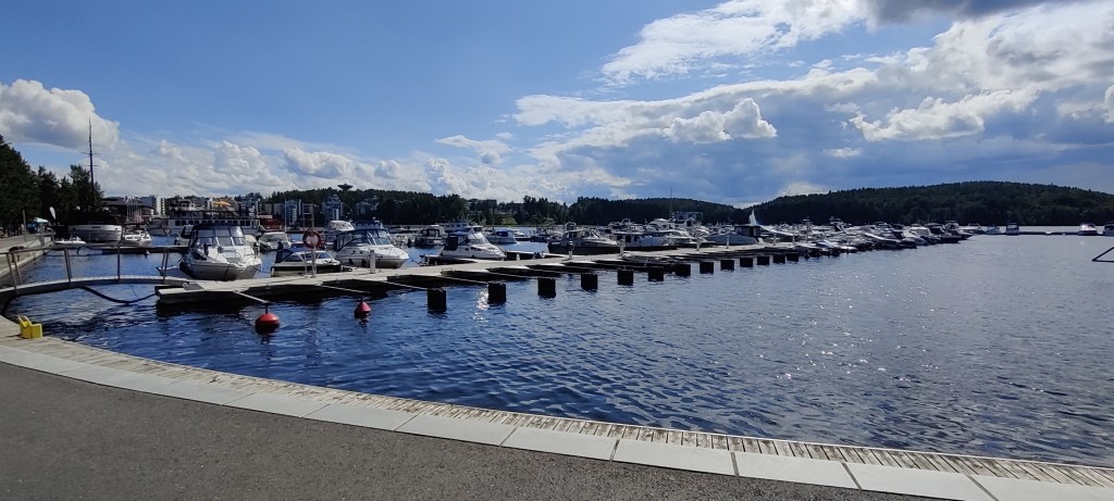 Kuvassa satama, jossa pitkää järveen työntyvää laituria reunustaa melkoinen määrä erilaisia veneitä. Taustalla näkyy Kuokkalan metsikköä ja vesitorni. Ilma on aurinkoinen, taivaalla on joitain valkoisia pilviä.