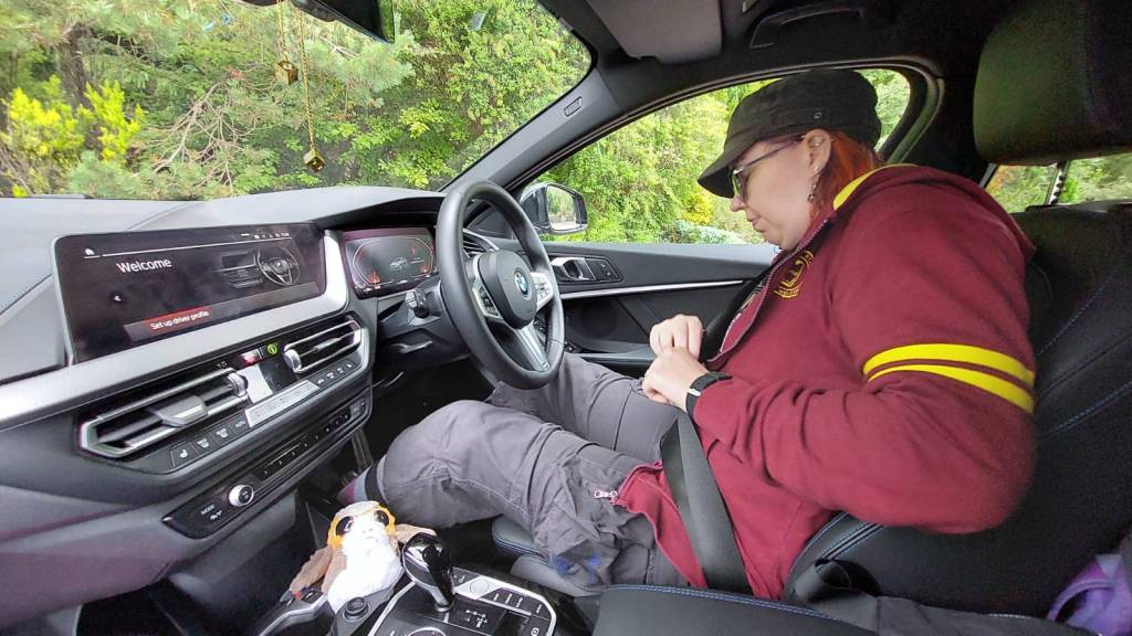 Pelkääjän paikalta otettu kuva kuskinpenkillä istuvasta Katjasta, joka säätää jotain turvavyön kanssa. Katjalla on lippis, aurinkolasit ja Tony Stark -huppari. Auton konsolinkolossa istuu pehmolelu-porg.