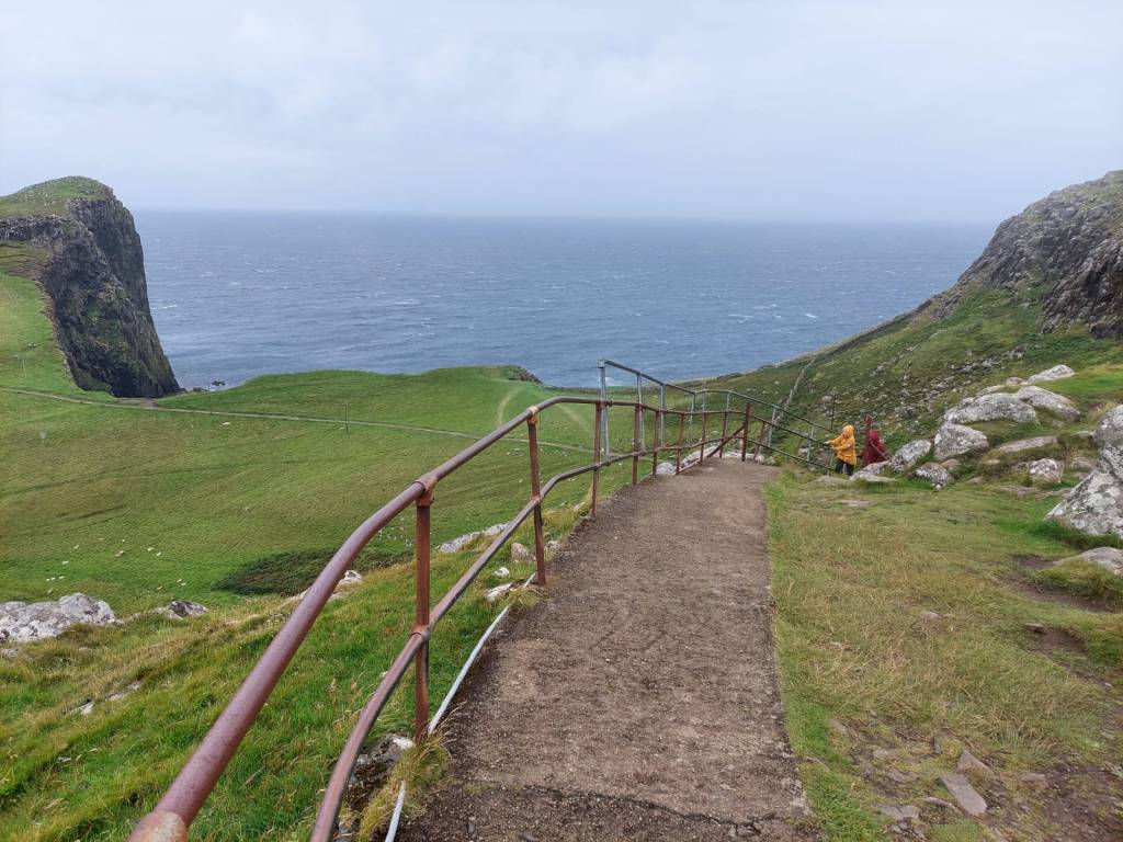 Laatoittua polkua reunustaa metallinen kaide. Polku katoaa jyrkkään rinteeseen, josta Taina ja Katja ovat kipuamassa pitäen tiukasti kaiteesta kiinni. Kuva on otettu korkealta, korkeuserot näkyvät. Vasemmassa reunassa erottuu kallio, taustalla tumma meri ja pilvinen taivas.