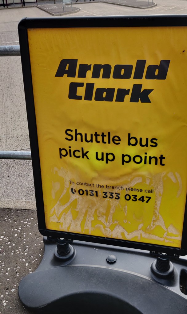 Keltainen mainoskyltti, jossa lukee mustalla "Arnold Clark, Shuttle bus pick up point" sekä puhelinnumero.