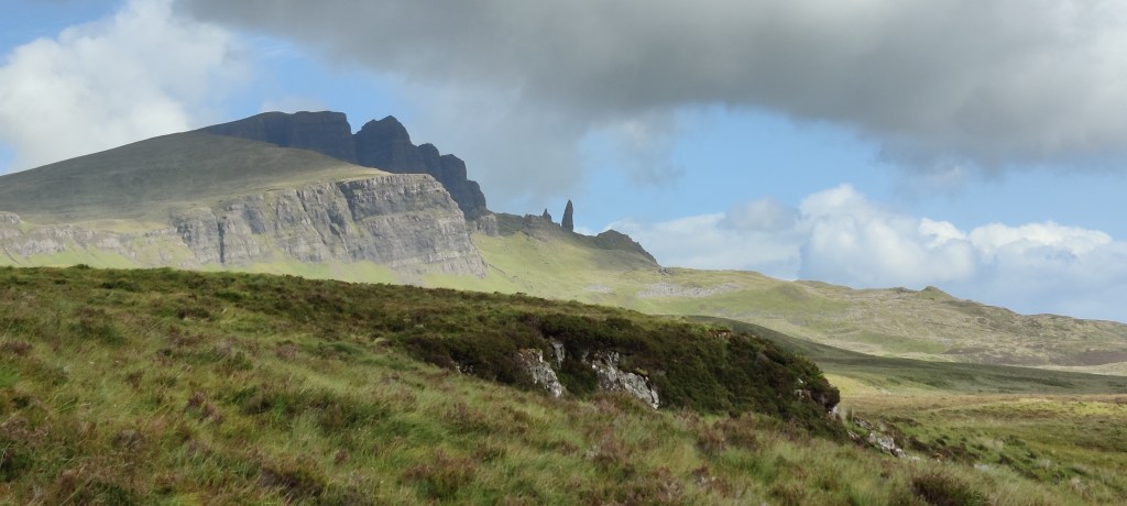 Karu maisemakuva heinikkoisesta rinteestä. Kaukana kohoaa tasainen kallio, jonka takana tummempia vuorenhuippuja. Niiden oikealla puolella kohoaa yksinäinen kivipylväs. Pilvet tekevät varjoja maastoon, taivalla roikkuu haarmaa pilvi matalalla.