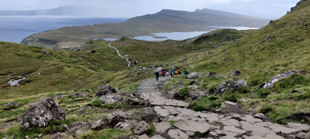 Rinteestä alaspäin otettu kuva, jossa kivistä koostettu polku muuttuu hiekkapoluksi alaspäin mennessä. Polulla kulkee kauempana ihmisiä värikkäissä takeissa. Polkua reunustaa suuria kiviä ja karua vihertävää maastoa. Rinteiden takaa kuultaa järvi ja meri ja vuoristoiset maastot.