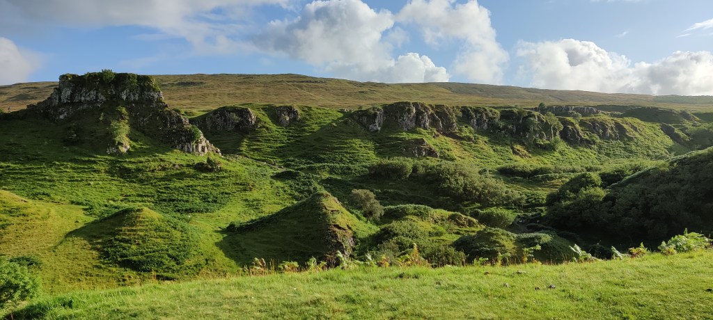 Vihreä, pieniä kumpuja täynnä oleva maisema. Taustalla kivisemmät, tasapäiset kukkulat kohoavat. Sää on aurinkoinen, taivaalla joitakin pilviä.
