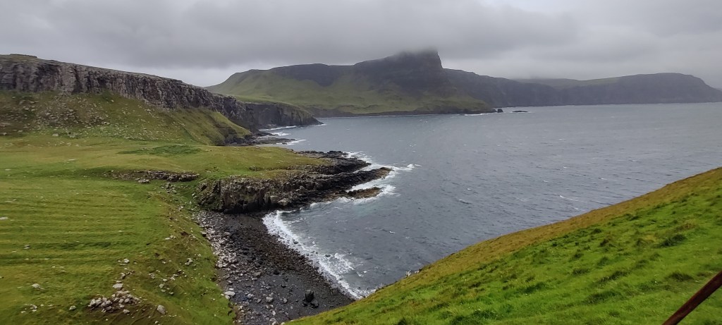 Rinteestä otettu kuva karusta rannikosta. Matalaruohikkoinen niitty on vihreä, ranta tumma ja kivikkoinen. Kiviset kalliot kohoavat rannasta. Taustalla näkyy lisää kallioita ja huippu, jonka harmaat pilvet peittävät. Vesi ja taivas ovat harmaita.