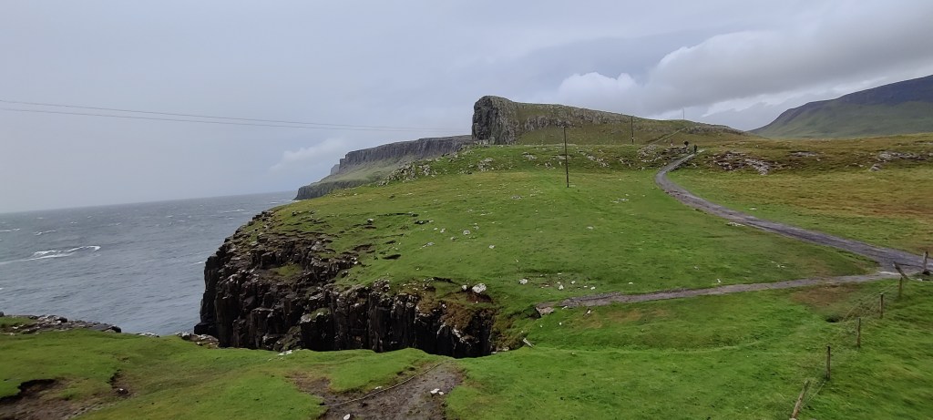 Vihreän nurmikon peittämä kallioranta, jonne vie kapea polku. Oikeassa reunassa erottuu aidanreuna. Kalliot ovat korkeita, merellä näkyy vaahtopäitä. Sähkötolpat kertovat sivistyksestä.