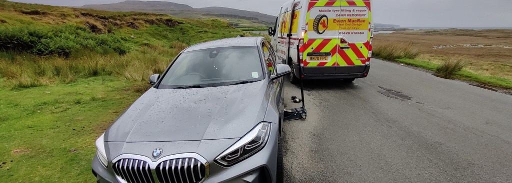Kuvassa tienposkessa seisova harmaa BMW, jonka alla seisoo musta tunkki. Takana on punakeltainen hälytysajoneuvo, jonka takaikkunassa on renkaan kuva. Taustalla näkyy numminen maisema.