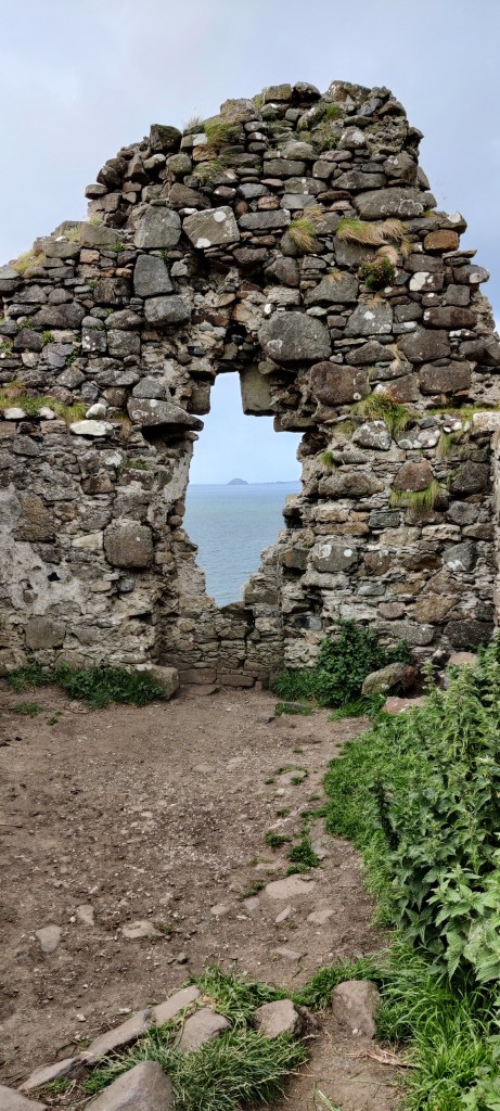 Kivinen raunioseinä, jonka keskellä on soikea ikkuna-aukko. Aukosta näkyy meri ja pieni saari. Etualalla on hiekkamaata ja nokkosia.