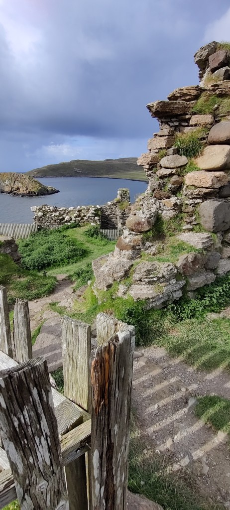 Pätkä raunioiden muuria, jota näkyy vielä kauempana lisää. Etualalla pala puista aitaa, jollaista näkyy kauempana muurin vierellä. Maasto on epätasaista ja ruohikkoista. Kaukana näkyy merenlahti ja kivikkoinen saari.