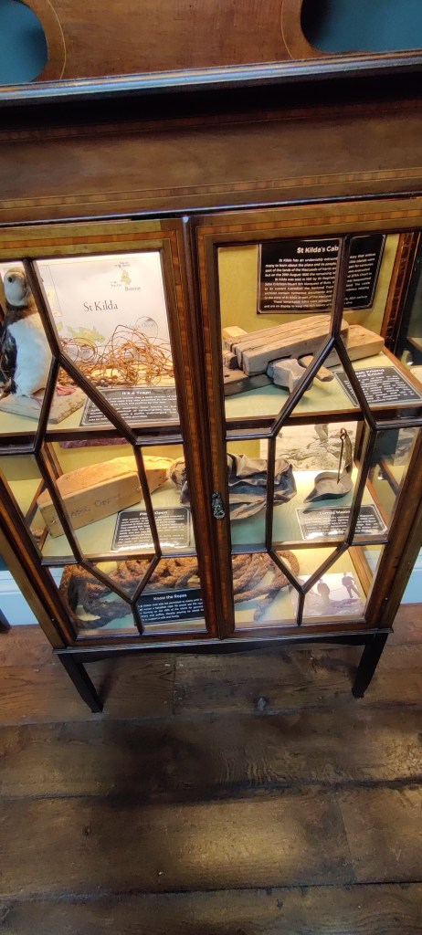 Ruskea vitriinikaappi, jossa on kolme hyllyä ja paljon erilaisia esineitä, mm. lunnipatsas.