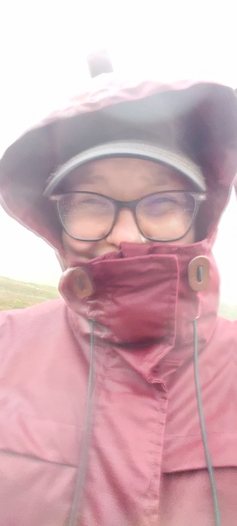 Selfie, jossa Katja on hautautunut vedenpitävään, hupulliseen takkiinsa. Hupun alta erottuu märät silmälasit ja sirrissä olevat silmät. Kuva on sumea ja epätarkka, takki näyttää litimärältä.