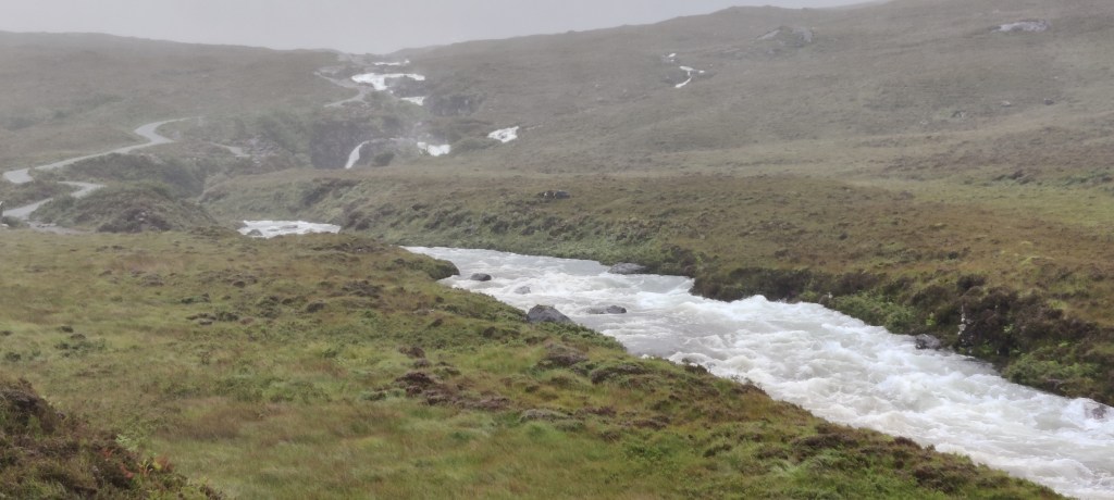 Utuisen sateinen kuva nummien keskellä virtaavasta joesta, joka vaahtoaa pieninä koskina. Joen vieressä kulkee mutkitteleva polku.