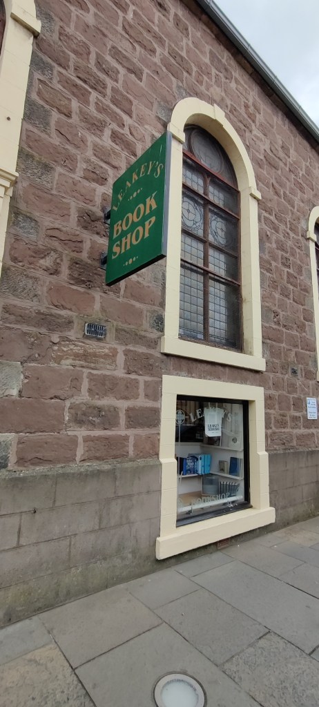 Kuva tiiliseinäisestä rakennuksesta, jossa on kaksi päällekkäistä ikkunaa. Ylempi on korkeampi, pyöreä yläkarmiltaan. Sen vieressä on vihreä kyltti, jossa lukee Leaky's Bookshop.