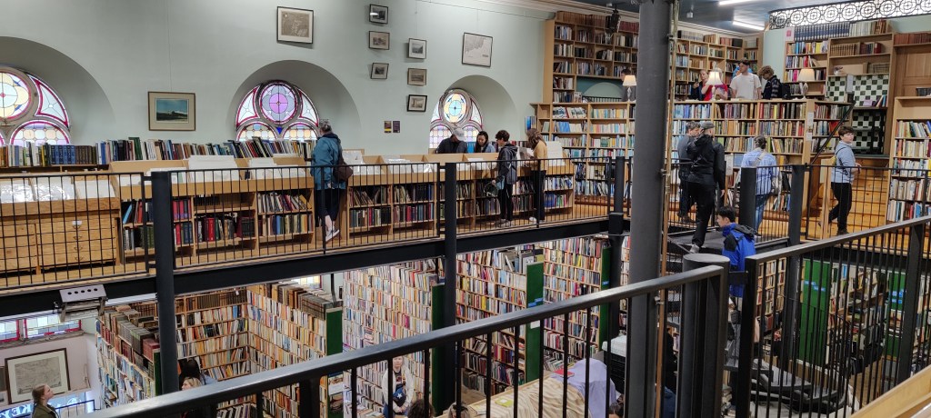 Kuva avoimesta sisätilasta, yläkertaa kiertää mustat metalliset kaiteet. Taustalla näkyy kirjahyllyjä ja ihmisiä selaamassa tarjontaa. Vastapäisellä seinällä on pyöreitä holviaukkoja, joissa on lasimaalauksin koristettuja pyöreitä ikkunoita.