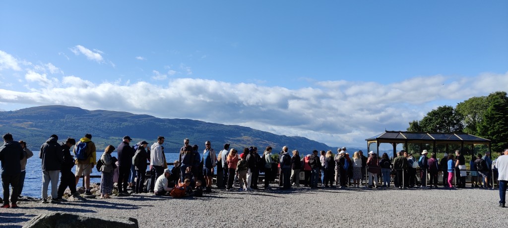 Aurinkoisessa säässä hiekkaisella rannalla oleva pitkä jono ihmisiä. Taustalla näkyy vettä ja vastarannan kukkulat.