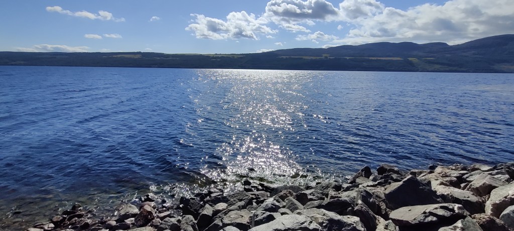 Kivikkoiselta rannalta otettu kuva järvelle. Vastarannan kumpuileva maasto näkyy. Järven pinta aaltoilee pienesti, auringonpaiste heijastuu vedestä.