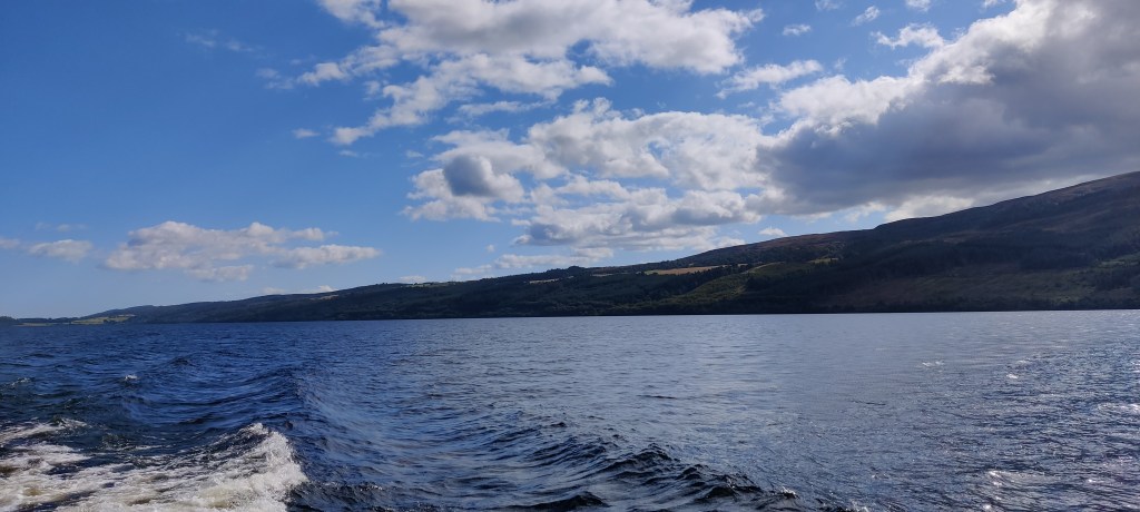 Veneestä otettu kuva kohti järvenselkää. Vastarannan kumpuileva maasto erottuu. Taivas on puolipilvinen. Veneen jättämät valkoiset mainingit näkyvät kuvan alareunassa.