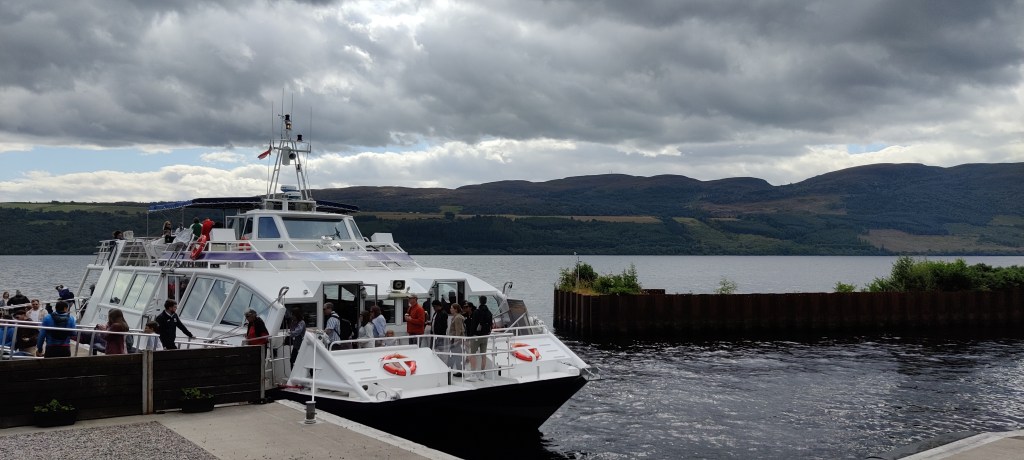 Laiturissa kiinni oleva valkoinen katamaraani. Ihmiset nousevat veneestä rantaan. Järven vastaranta näkyy, taivas on pilvinen.