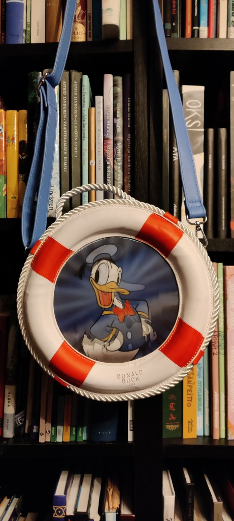 Kuvassa kirjahyllyn edessä roikkuva olkalaukku, joka näyttää pelastusrenkaalta, jonka keskellä on kuva Aku Ankasta. Tietyssä kulmassa Aku näyttää värikkäältä, modernilta Akulta, toisesta kulmasta mustavalkoiselta Akulta Akun ensimmäisestä animaatioelokuvasta.