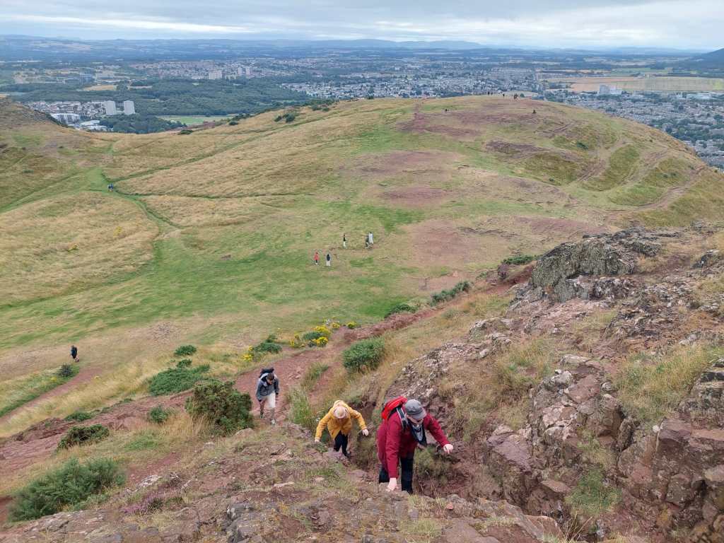 Kuva huipulta alas. Katja ja Taina nousevat kivikkoista rinnettä ylöspäin. Alla näkyy muutamia ihmisiä ruohikkoisella kukkulalla. Taustalla, kukkulan takana Edinburghin kaupunki.