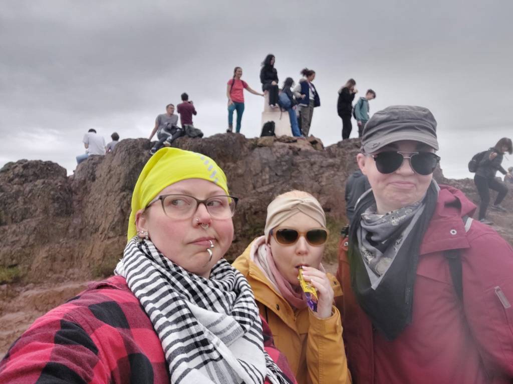 Selfie, jossa Hanna, Taina ja Katja ilmeilevät. Tainalla ja Katjalla on aurinkolasit. Taustalla kallioisen kukkulan huipulla seisoskelee joukko teinejä.