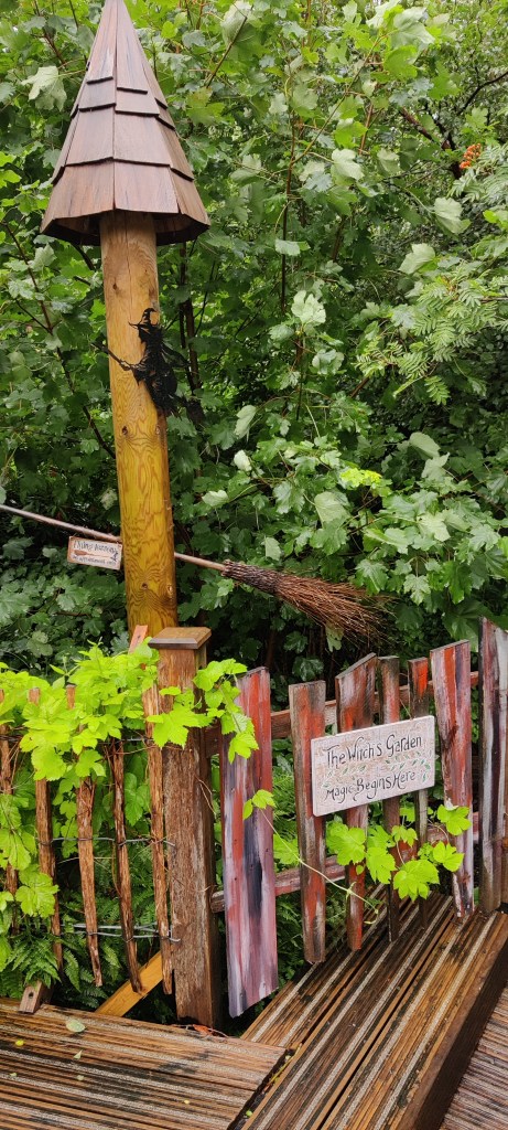 Puuaidassa kyltti, jossa lukee "The Witche's Garden. Magic begins here. Aidan takana pylväs, jossa loidan luuta ja musta noitahahmo