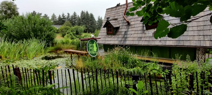 Matalan aidan takana vihreää, kasvustoa täynnä oleva pieni lampi ja vanha rakennus. Vihreässä kyltissä lukee Fairytale Distillary