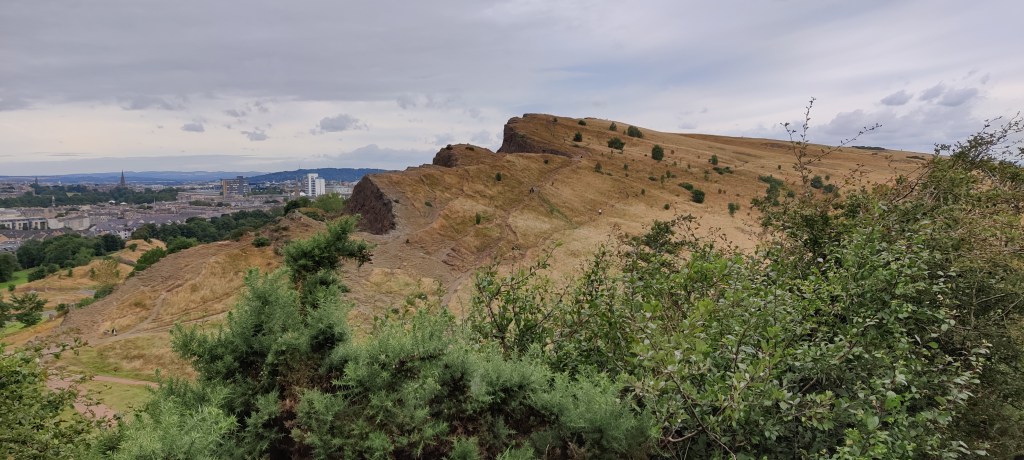 Maisemakuva, jossa Arthur's Seatin kukkula peittää osin Edinburghin kaupunkia. Kuva otettu ylhäältä, itse kukkulalta. Etualalla on pensaikkoa.