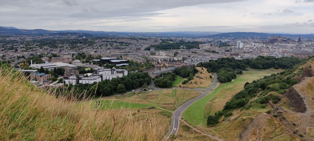 Kuva polulta alas: etualalla heinikkoa, alhaalla näkyy autotie, joka vie kaupunkiin. Taustalla Edinburghin kaupunki levittäytyy kaukaisuuteen.
