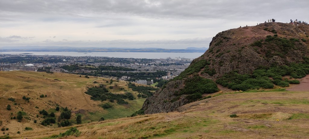 Maisemakuva kukkuloilta. Horisontissa näkyy Edinburghin kaupunki ja merenranta, etuoikealla Arthur's Seatin huippu, jolla seisoo joukko ihmisiä.