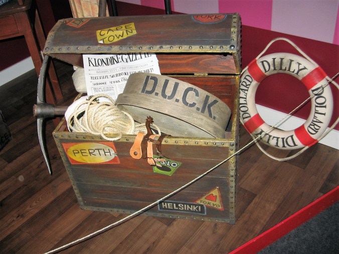 Kuvassa on näyttelyssä oleva puinen matkakirstu, joka on auki ja täynnä selakaista sälää. Siihen on liimattu eri kaupunkien (mm. Perth, Helsinki ja Tokio) tarroja. Siellä lukee myös isolla D.U.C.K. Vieressä on vanha pelastusrengas.