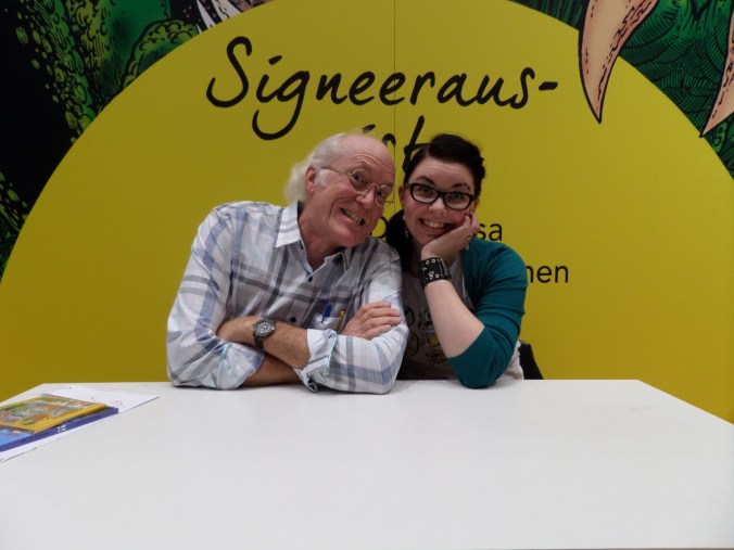 Katja istuu Don Rosan vieressä pöydän takana. Taustalla lukee signeeraus. Katja pitelee Donia käsivarresta. Kumpikin hymyilevät leveästi.