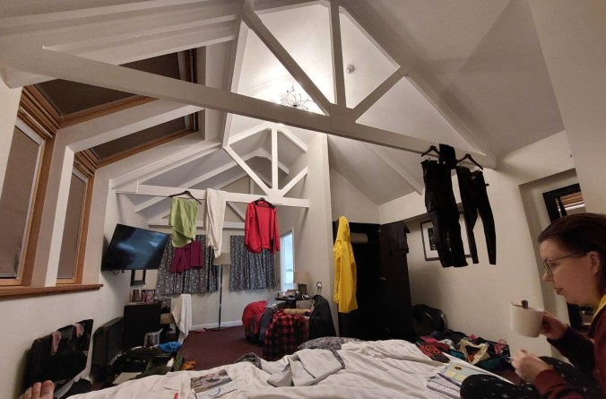 Sängystä päin otettu kuva suuresta hotellihuoneesta, jonka korkea katto erottuu hyvin. Katon olla on koristeellisia tukipalkkeja, joista roikkuu lukuisia vaatteita kuivumassa. Tavaroita on levällään ympäriinsä ja kuvan etureunassa näkyy Katja, joka pitelee valkoista kaakaomukia kädessään.