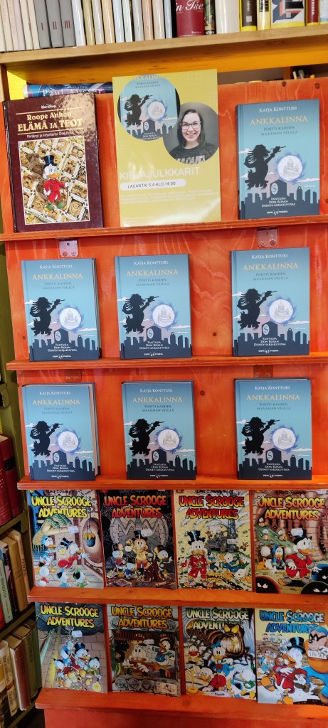 Näyttelyhylly, jossa useita kappaleita Katjan Ankkalinna-kirjaa esillä. Mukana myös Rosan Roope Ankan elämä ja teot, ja alahyllyillä lukuisia englanninkielisiä Disney-sarjakuvalehtiä, joiden kannet ovat Don Rosan käsialaa. Ylimmän hyllyn keskellä on mainos julkkareista.