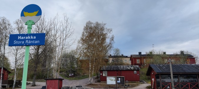 Maisemakuva rakennuksista, etualalla kyltti, jossa lukee Harakka, Stora Räntan ja sen päällä Helsingin kaupungin vaakunan vene.