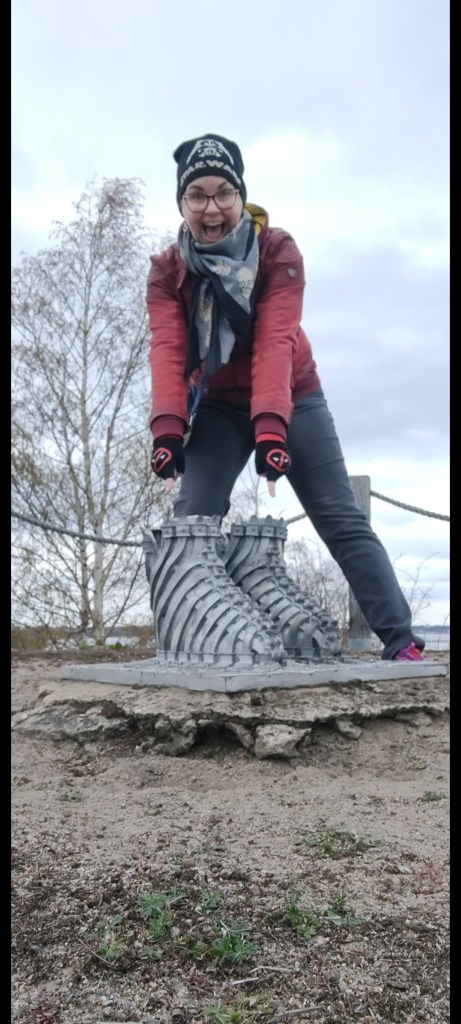 Katja seisoo veistoksen takana ja osoittaa sitä etusormillaan innostuneena, suu auki. Katjalla on punainen nahkatakki, Star Wars -pipo ja Deadpool-kynsikkäät.