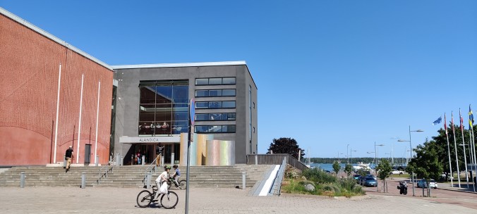 Kaupunkimaisemakuva, jonka vasemmassa reunassa on laatikkomainen kongressirakennus, jonka vasen seinusta on terrakotan oranssi. Pääoven yläpuolella on runsaasti ikkunoita ja kyltti, jossa lukee ALANDICA. Sen edessä on kirjava taideteos. Taustalla oikealla näkyy katu, jonka varrella on pohjoismaisia lippuja sekä kaukana taustalla meri.