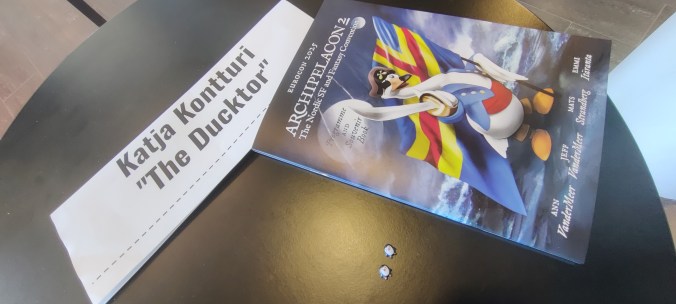 Pyöreällä pöydällä oleva nimikyltti, jossa lukee Katja Kontturi "The Ducktor". Sen vieressä on Archipelacon 2:n ohjelmalehtinen. Pöydältä erottuu myös kaksi pientä pingviinihahmoa.