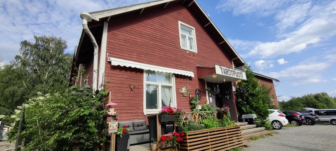 Punaisen talon pääty rauhallisessa pihapiirissä. Etualalla kukkalaatikoita ja pensas. Taaempana näkyy parkkeerattuja autoja.
Oven yläpuolen katoksessa lukee Tarotpuoti.