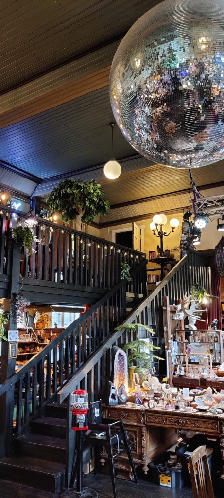 Kuva kaupan sisältä: siinä näkyy ruskean kirjoituspöydän päälle olevia kristalleja ja muita kiviä. Pöydän vierestä nousee portaat yläkerran parvelle. Katossa roikkuu jättimäinen discopallo.