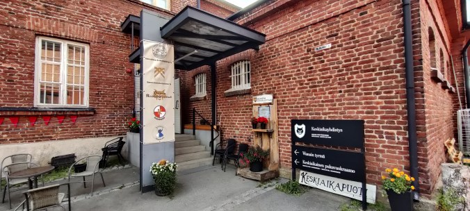 Tiilirakennuksen nurkkaus, jossa rappuset ja ovi sisälle. Kyltit maiostava Keskiaikayhdistystä ja Keskiaikapuotia.