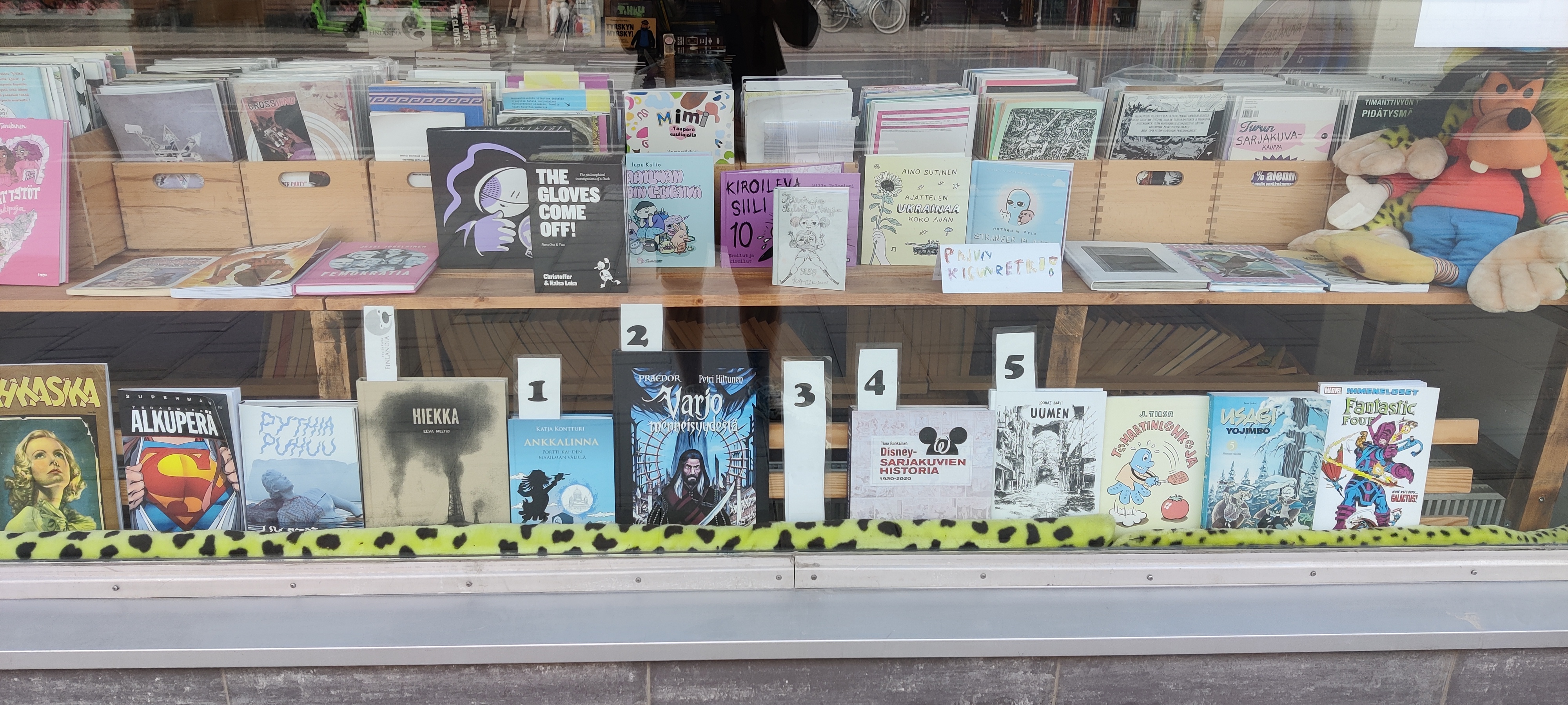 Kuva kirjakaupan näyteikkunasta, jossa on esillä lukuisia sarjakuvia. Etualalla on joukko teoksia, jotka on numeroitu yhdestä viiteen, joukossa muun muassa Petri Hiltusen uusien Praedor-sarjakuva. Ykkösenä on kuitenkin Katja Kontturin Don Rosa -kirja.