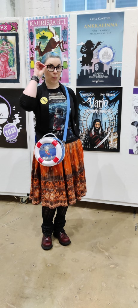 Kokovartalokuva Katjasta, joka poseeraa Aku-laukun kanssa oman kirjansa mainosjulisteen edessä.