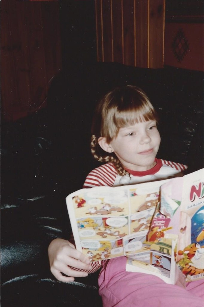 Vanha skannattu valokuva, jossa lettipäinen Katja lukee keskittyneesti mustalla nahkasohvalla Nalle Puh -sarjakuvalehteä.